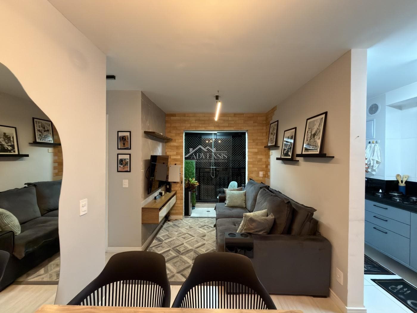 Sala de estar com sofá cinza, parede de tijolinhos aparentes e vista para a varanda com fechamento de vidro.