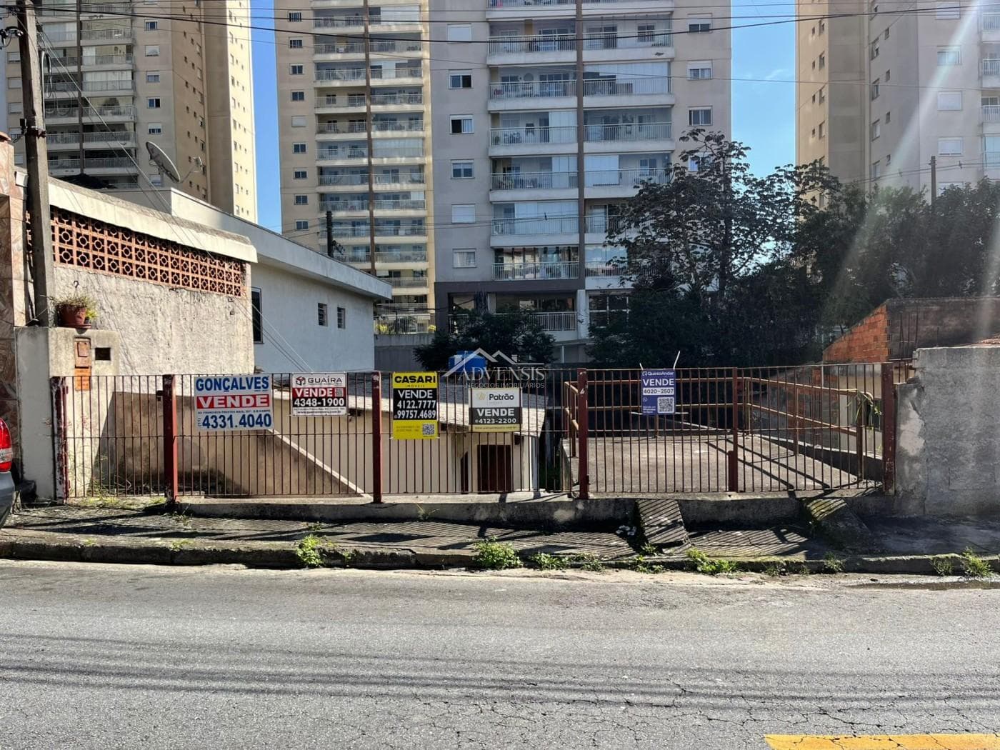 Fachada externa e portão de entrada com placas de imobiliárias e edifícios ao fundo
